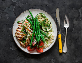 Delicious balanced lunch - grilled chicken fillet, orzo, string beans, zucchini and tomatoes on a dark background, top view