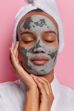 Verical close up of calm relaxed European woman with closed eyes doing cosmetology procedure applying face grey natural mask to keep skin fresh weaing white robe and towel standing isolated in centre