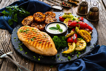 Grilled chicken breast, baked potatoes and barbecued  vegetables on wooden table
