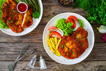 Breaded seared pork chop with fresh vegetable salad and French fries on wooden table
