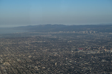 An aerial view over Los Angeles, showing its decentralized, sprawling urban planning with a few clusters of high-rise buildings. In the distance is the Pacific Ocean and some mountains beyond that.