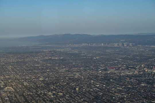 An aerial view over Los Angeles, showing its decentralized, sprawling urban planning with a few clusters of high-rise buildings. In the distance is the Pacific Ocean and some mountains beyond that.