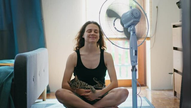 A young Caucasian woman holding a cute tabby cat in her arms while cooling down and relaxing in front of an electric fan at home during hot weather.