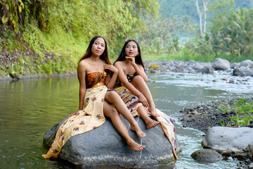 Two young beautiful Asian women sitting on a rock in a river.