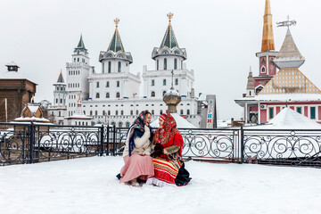 Obraz premium Two beautiful Slavic women in national traditional Slavic costumes on the roof of a wooden tower against the backdrop of a snow-covered city. Pancake day