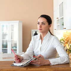 Beautiful young woman sitting at the table in the kitchen and taking notes
