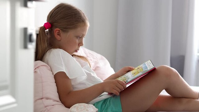 The child is reading. Girl sitting on the bed in the bedroom reads an interesting book. Portrait of a beautiful little girl reading a book sitting on a comfortable bed in the bedroom at home.