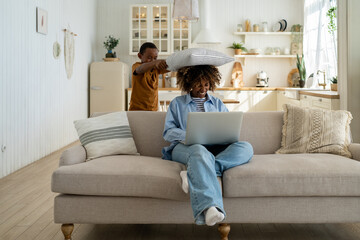 Pleased playful African American child boy pillow hit to surprised head mom busy work online shopping while sit with laptop on sofa. Happy family time mother and son together fun game activity at home