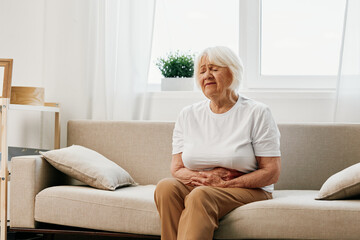 Elderly woman severe abdominal pain sitting on the sofa, health problems in old age, poor quality of life. Grandmother with gray hair holding her stomach, poisoning, problems with stool.