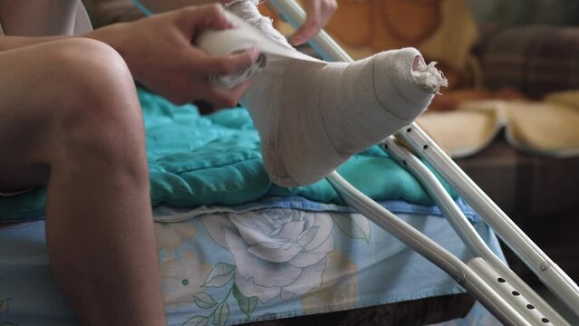 a man rewinds a sore leg with a plaster cast with an elastic bandage sitting sobbing crutches the concept of fractures and injuries, rehabilitation