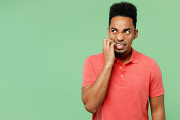 Young confused mistaken sad man of African American ethnicity wearing pink t-shirt look aside biting nails fingers isolated on plain pastel light green background studio portrait. Lifestyle concept.