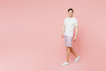 Fototapeta na wymiar Full body side view young caucasian man wearing white t-shirt casual clothes walking going strolling looking camera isolated on plain pastel light pink background studio portrait. Lifestyle concept.