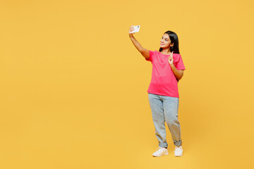 Full body young happy Indian woman wear pink t-shirt casual clothes doing selfie shot on mobile cell phone post photo on social network isolated on plain yellow background studio. Lifestyle concept