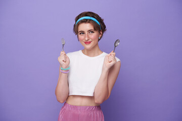 Portrait beautiful smiling young hungry woman holding spoon and fork isolated on purple background.