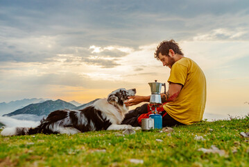 white man and young mountaineer sitting next to his border collie breed dog while preparing coffee...