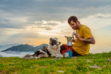Young white man with his border collie dog preparing a coffee on a camping stove at sunrise in the mountains. Hiking and adventure. travel with pet - obrazy, fototapety, plakaty