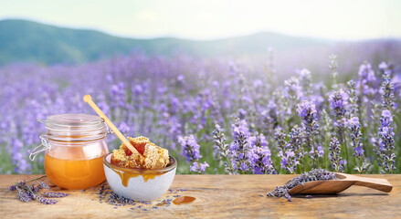 Lavender honey in a jar with honeycombs in a bowl and a scoop with dried lavender flowers on a wooden table. Mountain landscape with hills and lavender field in soft focus with bee products in front.