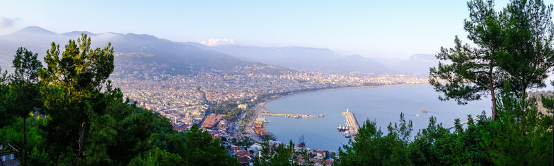 Fototapeta na wymiar Enjoy the stunning view of Alanya from the observation deck, with lush trees, blue sea, and the iconic Lighthouse and Port of Alanya. The rocky peninsula adds a dramatic touch to the panoramic scene.