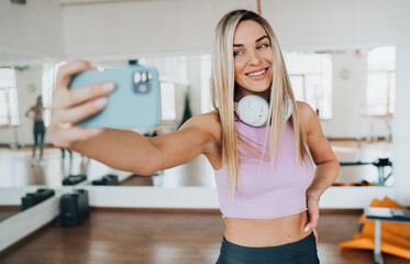 Beautiful fit European young woman in sportswear makes selfie at break of workout toothy smiles at gym. Excited blonde girl talks by phone via internet makes video call at fitness club.