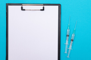 Fototapeta na wymiar Vaccination, Immunology or Revaccination Concept - Two Medical Syringe Lying on Blue Table in Doctor's Office in a Hospital or Clinic. Black Clipboard with Sheet of Paper - Mock Up with Copy Space