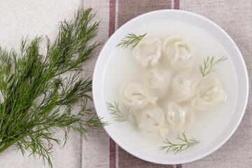 Bouillon with homemade pelmeni and fresh dill. Soup with dumplings and herbs on tablecloth. Top view
