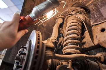 A car mechanic inspects springs, shock absorbers and suspension with a flashlight. Car on a repair stand. Technical service station for car. - 626546383
