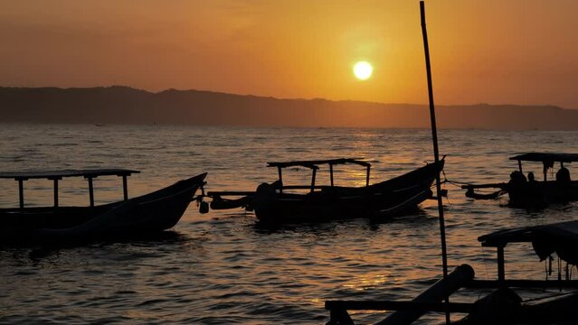 A silhouetted image of fishing boats moored on the shore of Pangandaran and rocked by the waves as the rising sun appears round above the skyline. Slow motion medium shot and handheld.