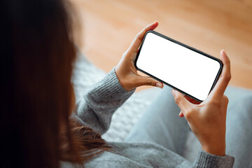 Closeup - Happy young woman holding mobile smartphone with blank white screen background while resting on the sofa in living room at home. Watching movies on the phone.