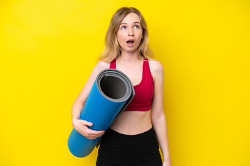 Young sport caucasian woman going to yoga classes while holding a mat isolated on yellow background looking up and with surprised expression