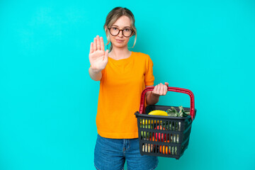 Blonde English young girl holding a shopping basket full of food isolated on blue background making...