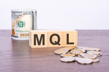 top view of wooden cubes with text MQL over US dollar banknotes and coins on a brown wooden background