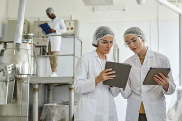 Waist up portrait of two young women wearing lab coats and using digital tablet in workshop of...