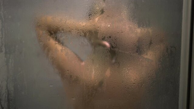 A young woman is washing herself in the shower. Misted shower glass