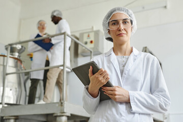 Waist up portrait of female worker wearing lab coat looking at camera in factory workshop, copy...
