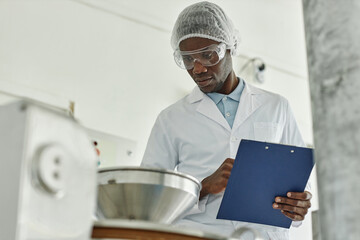 Portrait of black young man wearing lab coat and holding clipboard while overseeing production at food factory, copy space