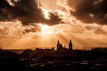 The setting sun shines through the clouds and colors everything orange. In the foreground the old town of Valletta