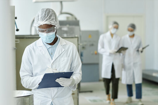 Waist up portrait of young black man wearing mask and holding clipboard looking in tanks at factory workshop while overseeing production, copy space