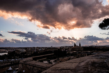 The setting sun shines through the clouds and colors everything orange. In the foreground the old town of Valletta
