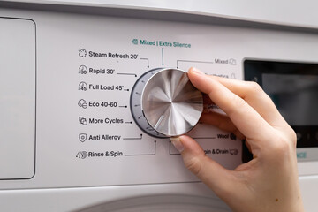 Woman hand choosing program on automatic washing machine for laundry at home. Turning knob on...