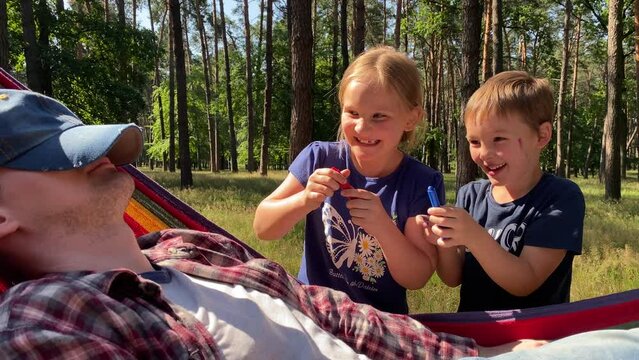 Family vacation in beautiful forest on sunny day. Cheerful kids sneakily paint father face as he naps in hammock in the woods. Family bonding time together while recreation in the great outdoors