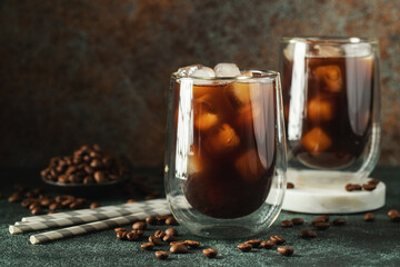 Ice coffee in a tall glass with cream poured over, ice cubes and beans on a dark concrete table.
