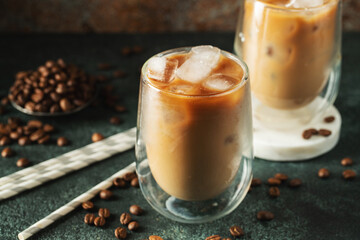 Ice coffee in a tall glass with cream poured over, ice cubes and beans on a dark concrete table.