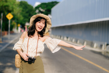 Portrait of asian young woman traveler with weaving hat and basket and a camera waving hand to friend by the street. Journey trip lifestyle, world travel explorer or Asia summer tourism concept.