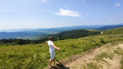 Fototapeta premium Young man standing with raised hands on top of a mountain and enjoying the view of the valley
