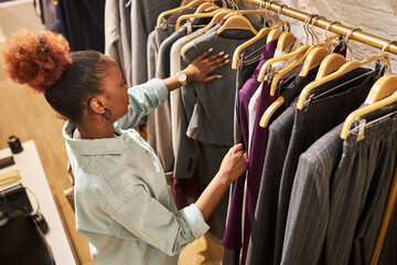 High angle portrait of African American young woman browsing clothes on rack in clothing store,...
