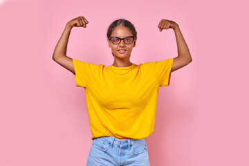 Young happy strong Indian woman in strongman pose shows muscles on arms to demonstrate self-confidence and readiness to achieve goals dressed in casual summer clothes stands on pink background