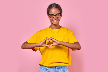 Young kind romantic Indian woman teenager makes heart shape from fingers as sign of love for boyfriend and hope for long relationship with subsequent wedding stands on pink studio background.