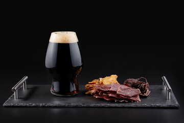 A tray with a glass of dark beer and dried meat on a dark background. Front view