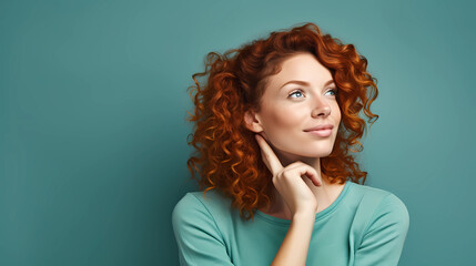 Headshot portrait of thoughtful pensive young ginger woman with curly hair holding finger behind ear looking upward against turquoise studio wall background with copy space for text advertisement 