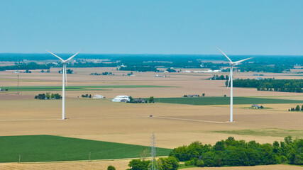 Fototapeta na wymiar Aerial two wind turbines in empty field with black truck on maintenance road and houses in distance
